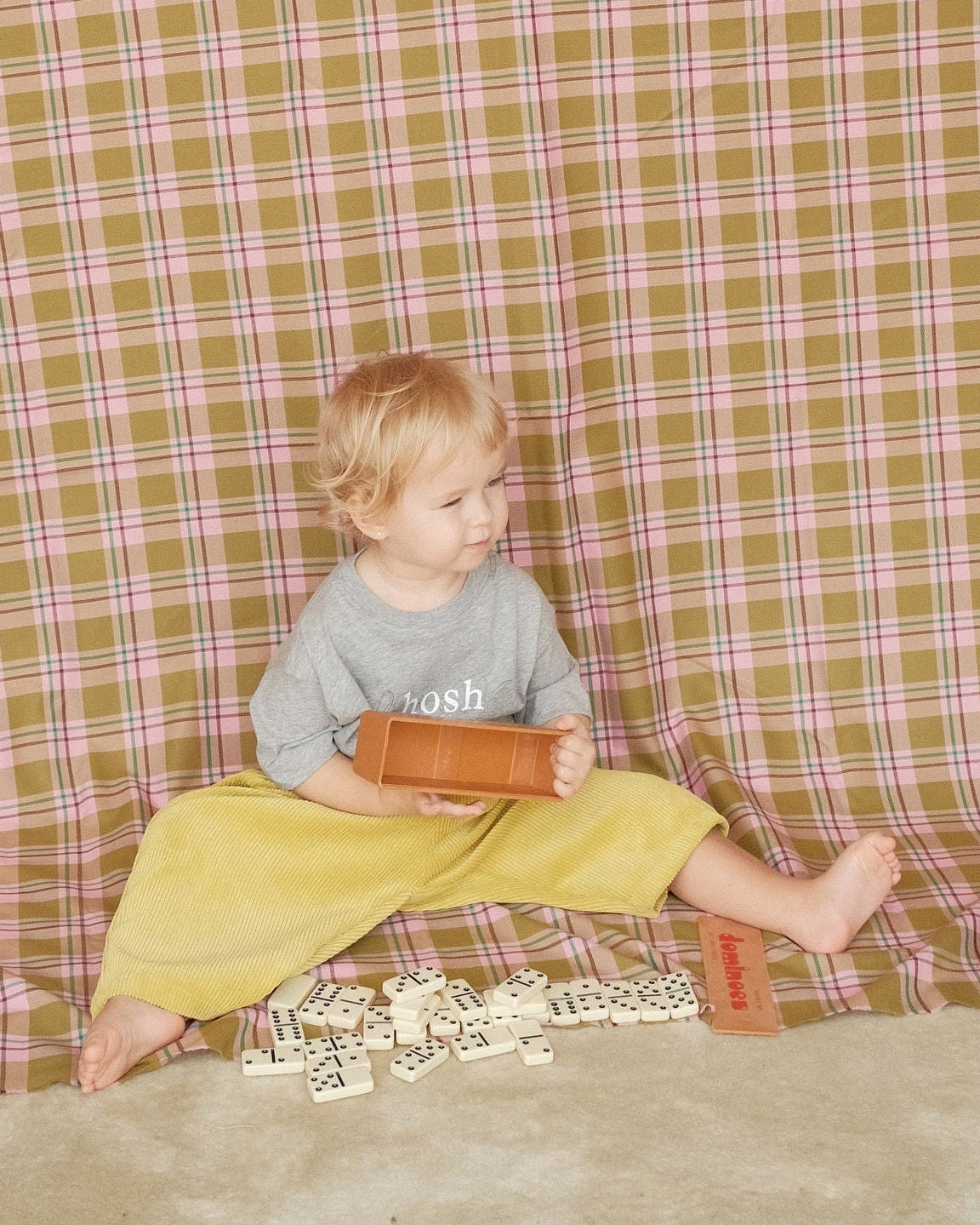 Child sitting on the floor with a checkered tablecloth draped over them, in a room with a wooden cabinet and air conditioner.