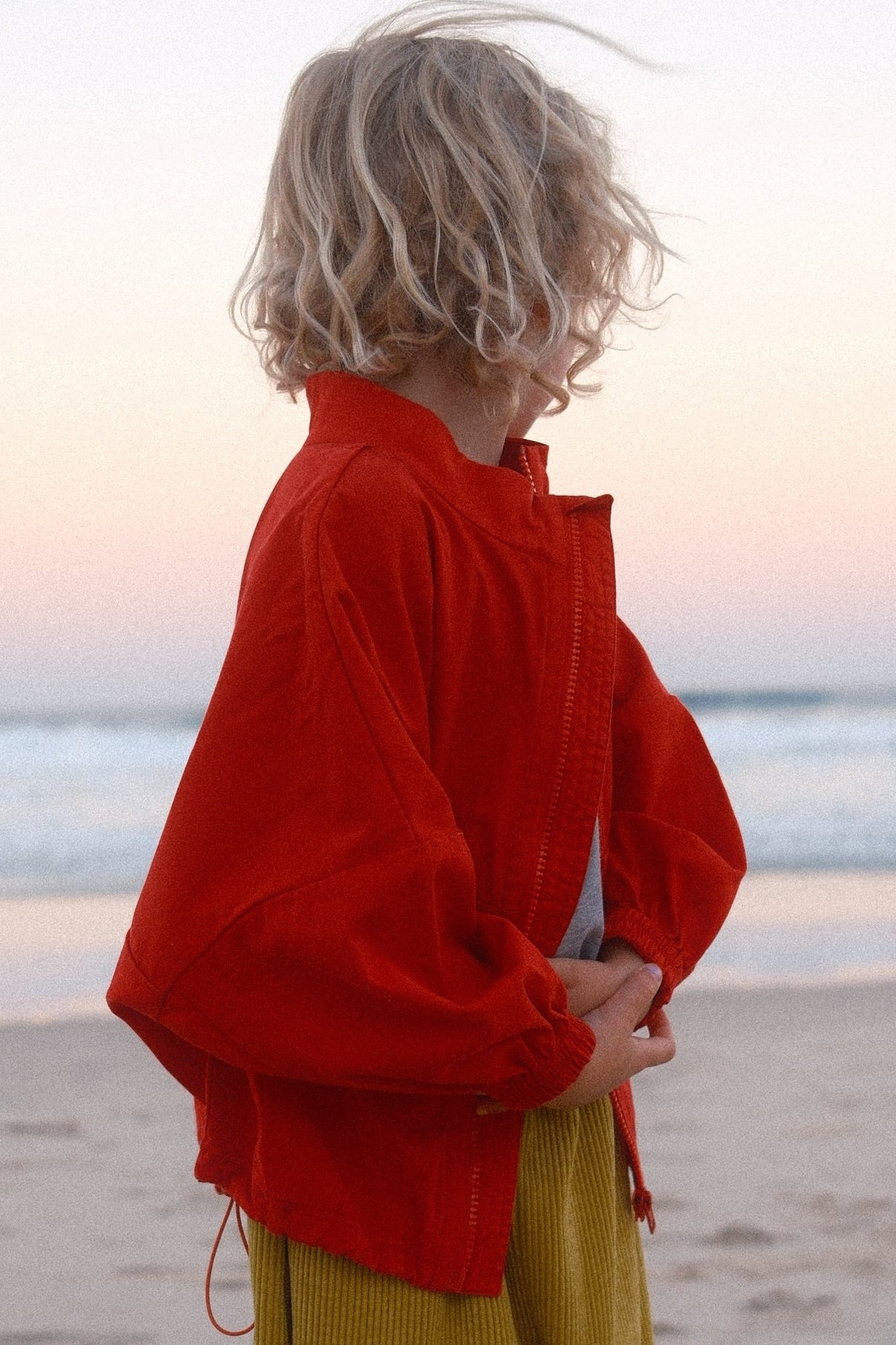 Person in red top and green pants standing on a beach with ocean in the background