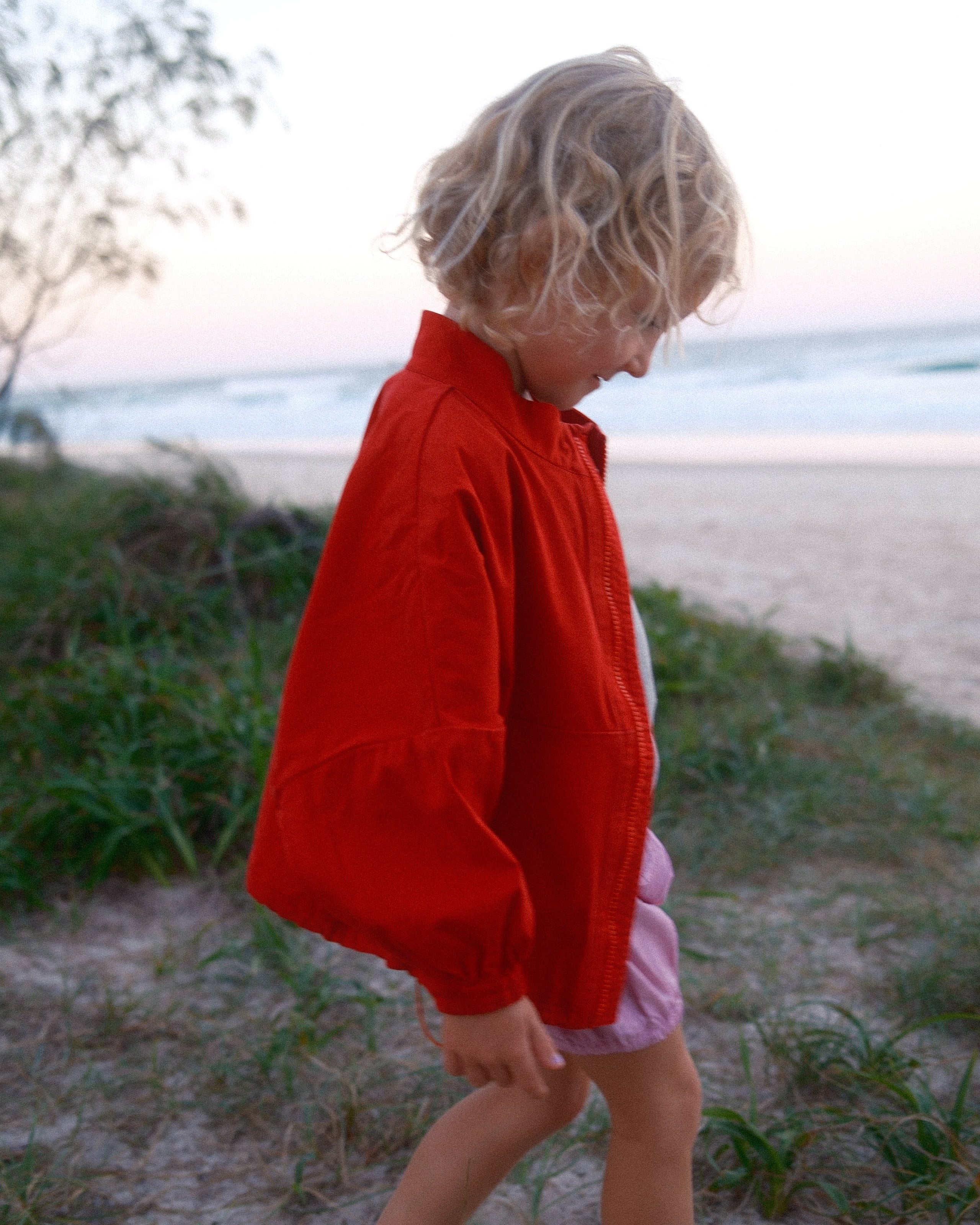 Child in a red shirt and pink shorts standing on a sandy path with grass.