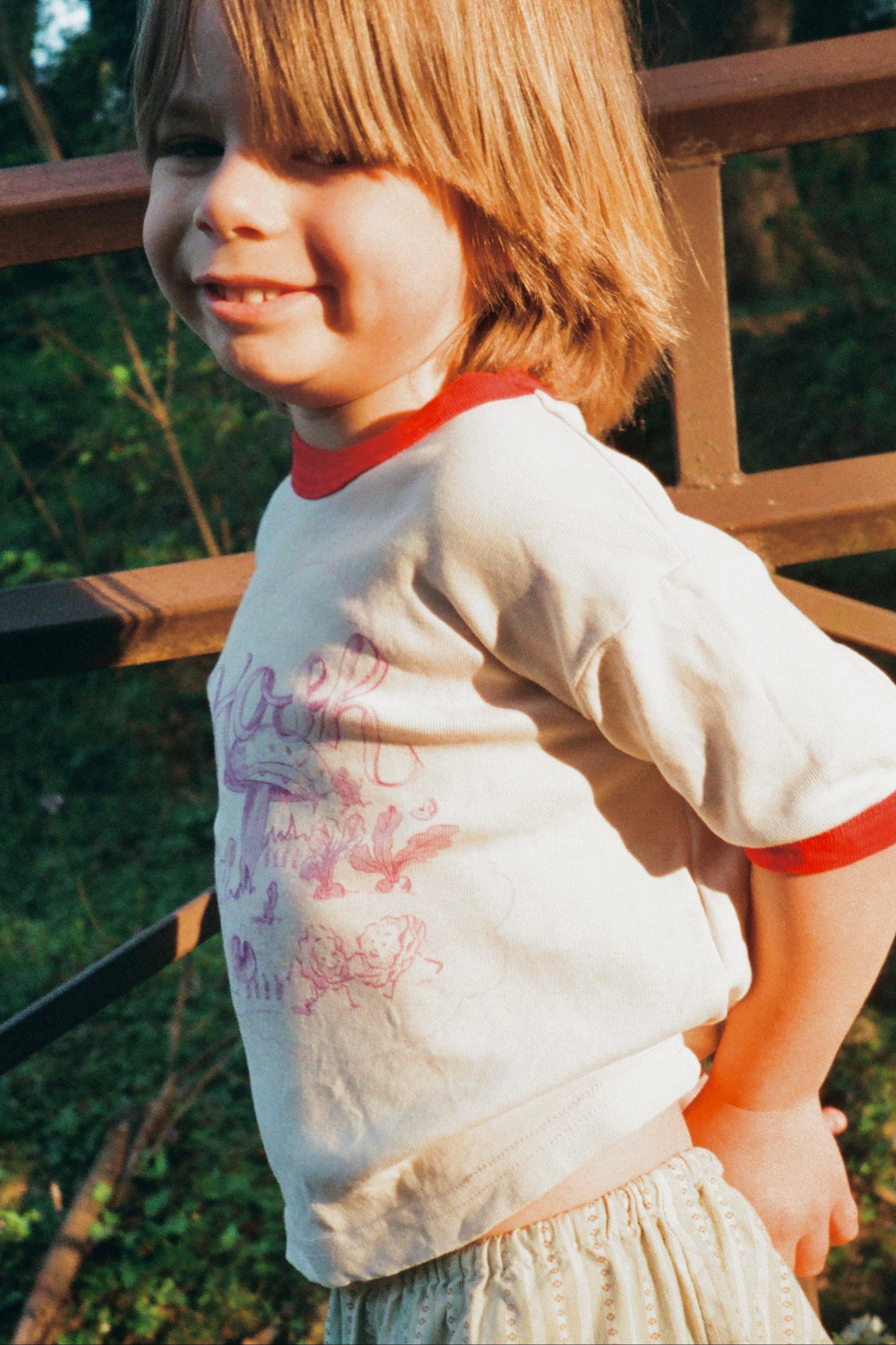 Child wearing a white shirt with red trim and beige pants standing outdoors near a wooden fence.