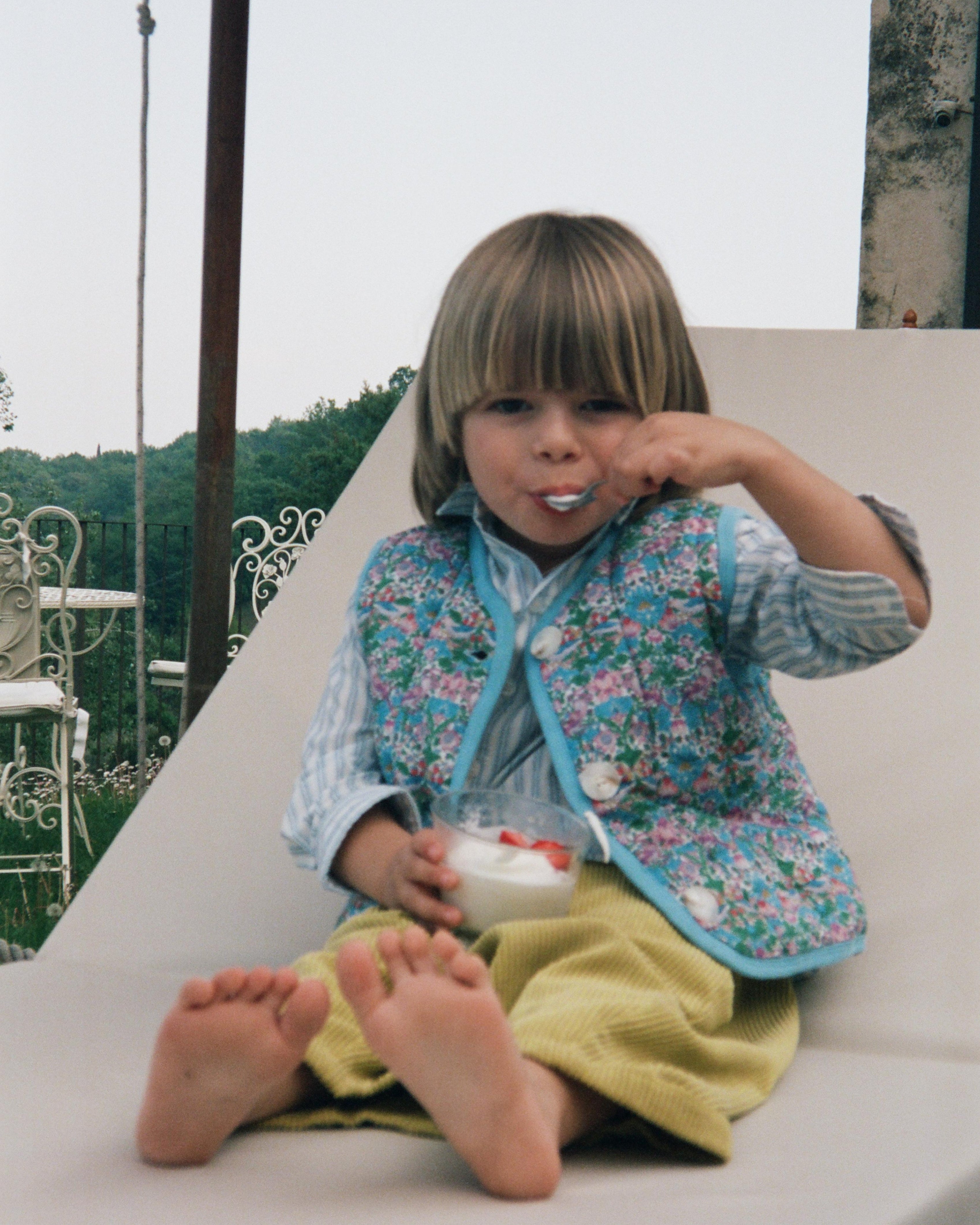 Child sitting on a white chair outdoors, eating from a bowl.