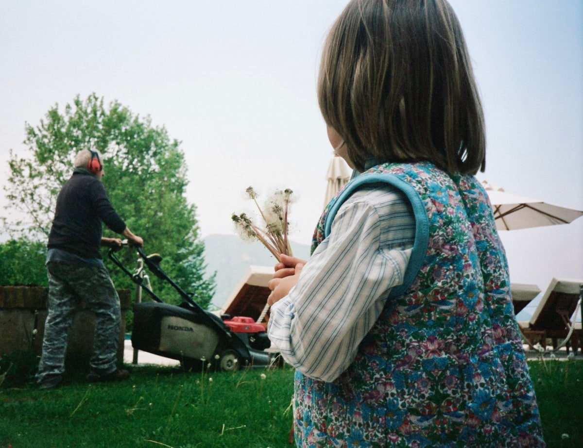Person mowing a lawn with another person observing, set against a backdrop of trees and open sky.