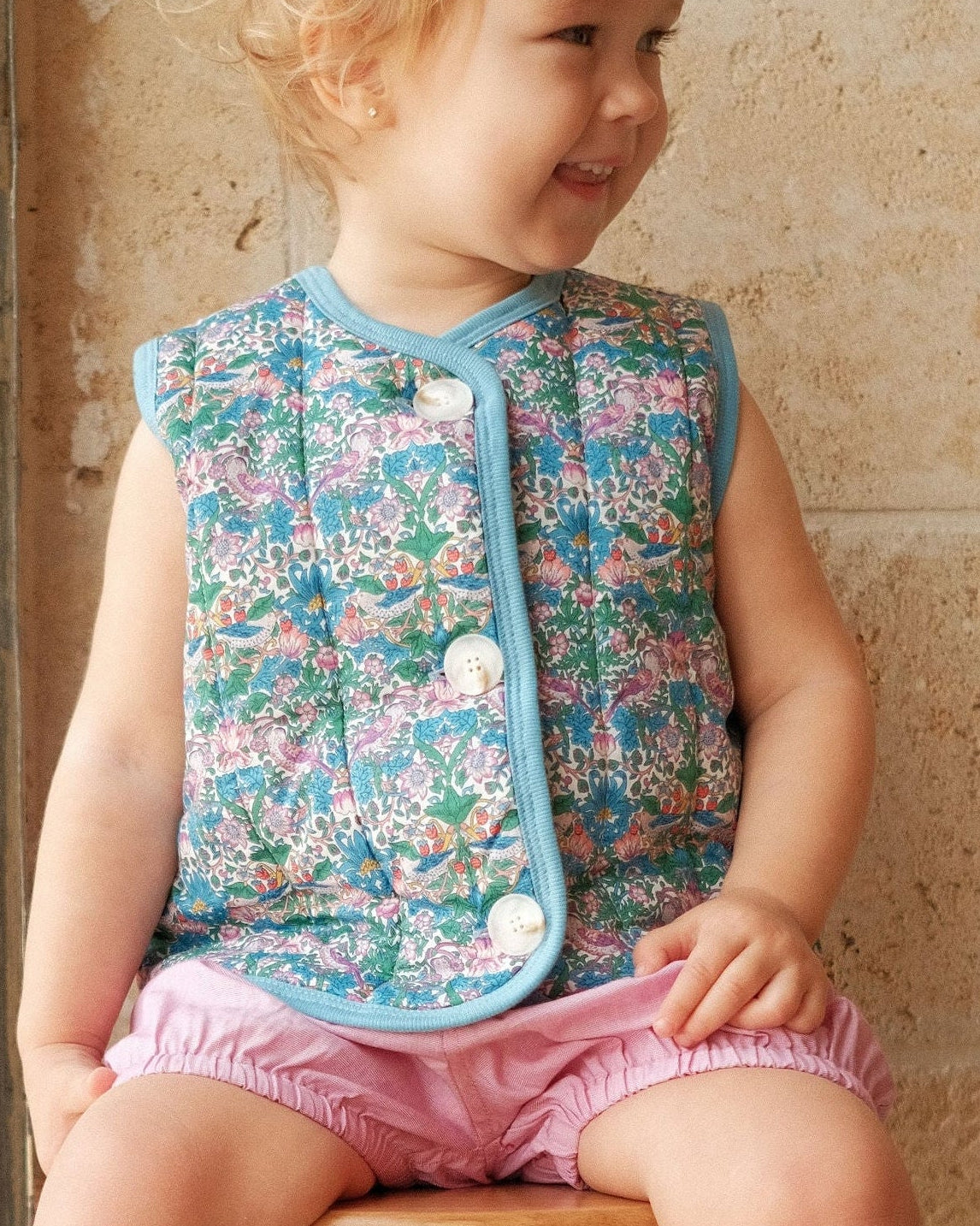 Child sitting on a stool wearing a floral patterned vest and pink shorts against a stone wall.