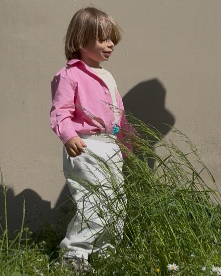 Child in a pink shirt and white pants standing in grass against a beige wall