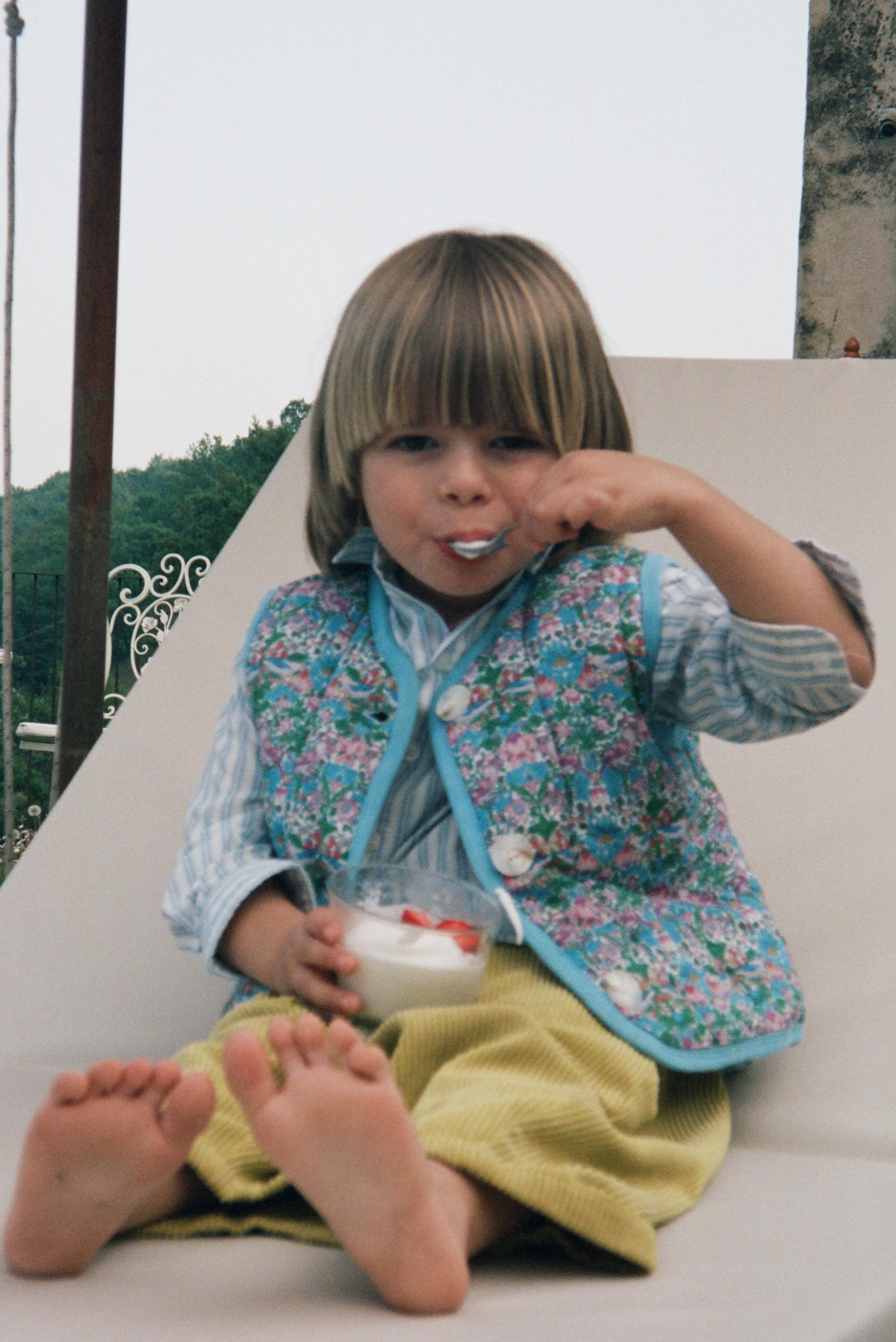 Child sitting on a white chair outdoors, eating from a bowl.
