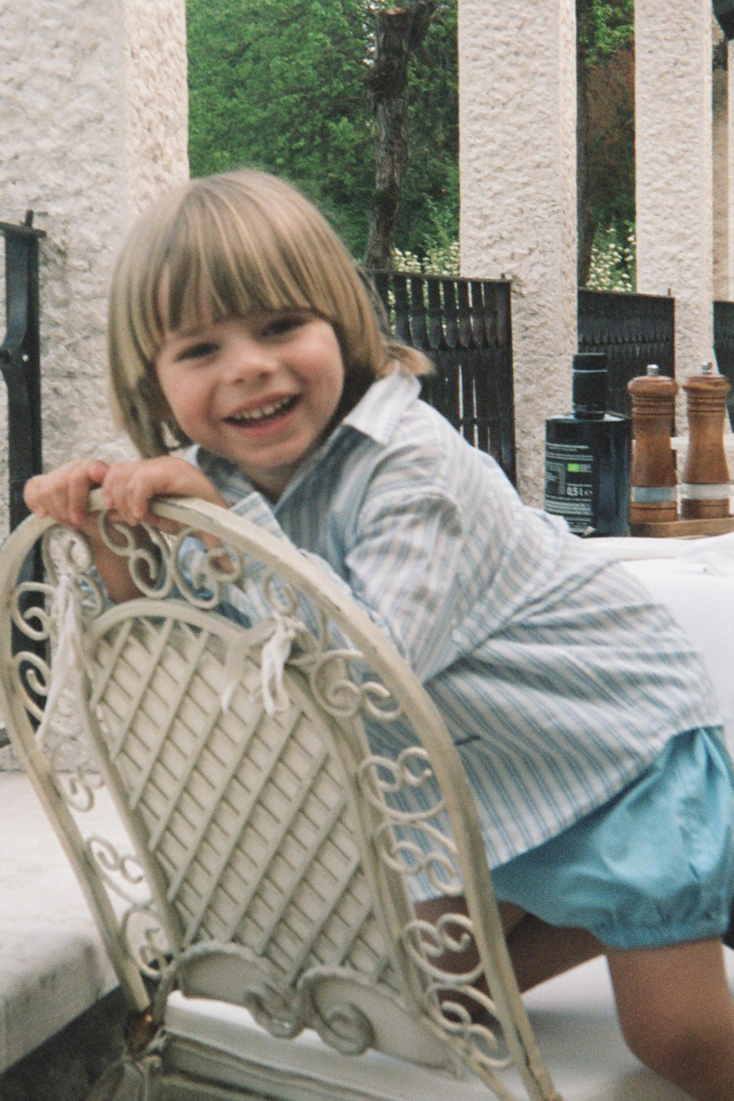 Child sitting on a white chair outdoors with trees and a building in the background